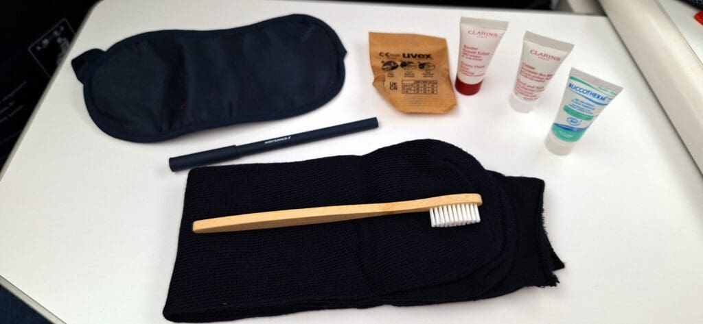 The image shows a collection of travel amenities on a white surface. Items include a black eye mask, a black pen with "Air France" written on it, a wooden toothbrush with white bristles, a pair of black socks, a brown paper packet, and three small tubes of skincare products.