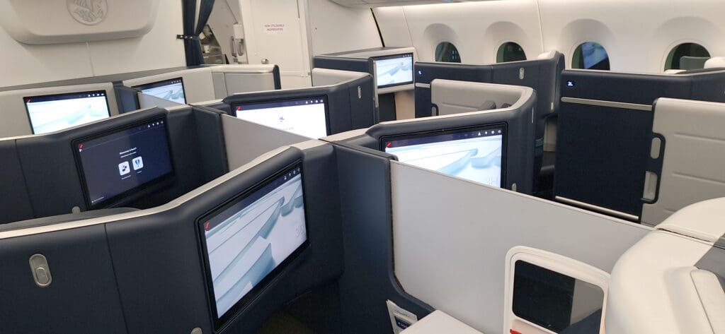 The image shows the interior of an airplane's business class cabin. The seats are arranged in a staggered configuration, each with a personal entertainment screen. The seats have high partitions for privacy, and the cabin has a modern design with a color scheme of dark blue and gray. There are windows along the side of the cabin.