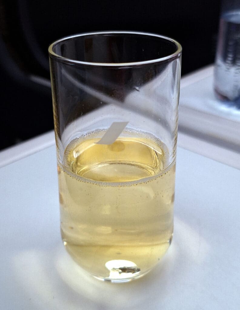 A clear glass filled with a light yellow liquid, possibly a beverage like apple juice or white wine, is placed on a white surface. There is a small white object, possibly a piece of paper or a stirrer, inside the glass. In the background, there is a blurred view of another object, possibly a bottle.
