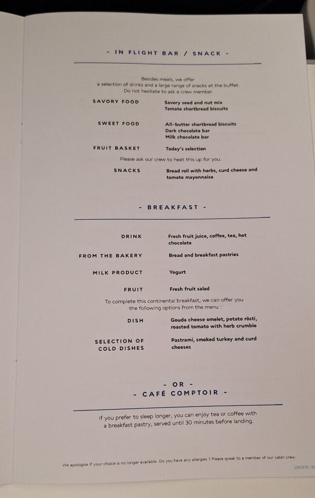 This image shows an in-flight menu with sections for snacks and breakfast. The snack options include savory food like a seed and nut mix, sweet food like chocolate bars, a fruit basket, and snacks such as a bread roll with herbs. The breakfast section offers drinks like juice and coffee, bakery items, yogurt, fruit salad, and dishes like a Gouda cheese omelet. There is also an option for a café service with tea or coffee and a pastry.