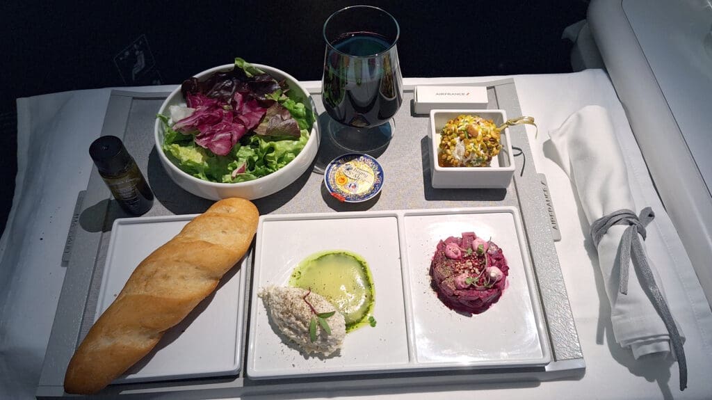 A meal served on an airplane tray includes a fresh green salad with mixed leaves, a small bottle of dressing, a glass of red wine, a piece of baguette, a cheese spread, and two plated dishes. One dish appears to be a creamy spread with herbs and oil, and the other is a beetroot salad with garnishes. There is also a small dessert topped with nuts, and a neatly rolled napkin with a tie. The tray is labeled with "Air France."
