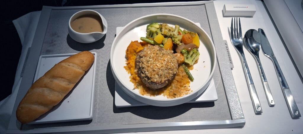 The image shows an airplane meal on a tray. There is a main dish consisting of a piece of meat topped with breadcrumbs, accompanied by mixed vegetables including broccoli and squash, and a side of mashed sweet potatoes. A small bowl of sauce is placed next to the main dish. On the left, there is a piece of bread on a separate plate. To the right, there is a set of silverware including a fork, spoon, and knife. The tray is labeled with "Air France."