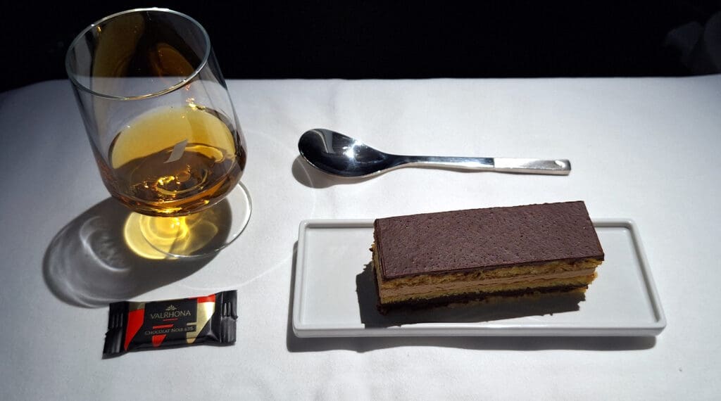 A glass of amber-colored liquid, possibly whiskey or brandy, is on a white tablecloth. Next to it is a silver spoon and a rectangular slice of layered chocolate dessert on a white rectangular plate. A small wrapped piece of Valrhona dark chocolate is placed nearby.