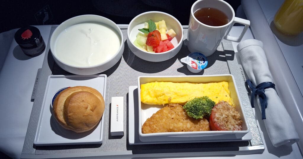 An in-flight meal on a tray, featuring a bowl of yogurt, a bowl of mixed fruit with a strawberry garnish, a cup of tea, a small jar of strawberry jam, and a wrapped butter portion. There is a plate with an omelet, hash brown, broccoli, and a tomato with breadcrumbs. A bread roll is on a separate plate, and a napkin is rolled with a blue tie.