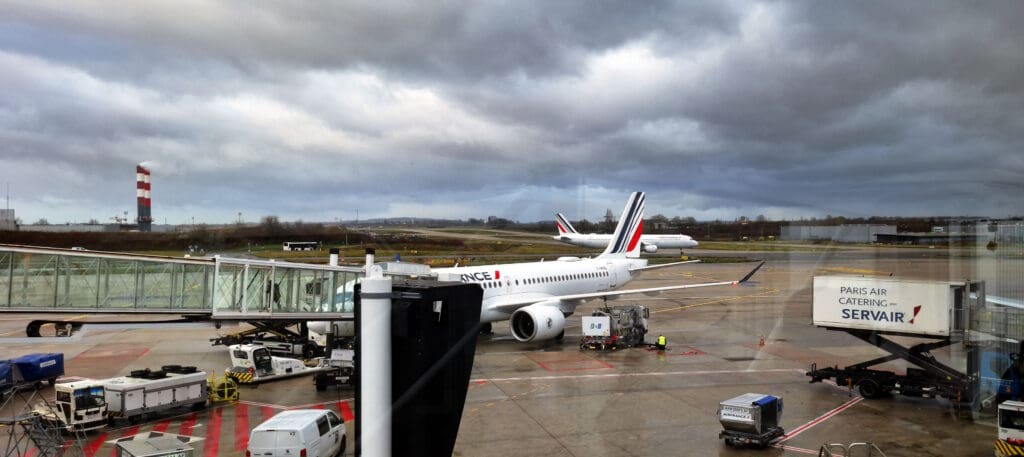 The image shows an airport scene with two airplanes on the tarmac. The foreground features an Air France plane parked near a jet bridge, with ground service vehicles and equipment nearby. In the background, another Air France plane is taxiing. The sky is overcast, and there is a red and white striped tower visible in the distance. A sign for "Paris Air Catering Servair" is also visible.