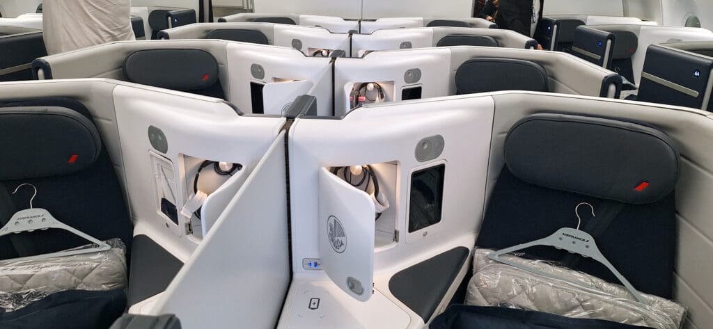 The image shows a luxurious airplane cabin with several business class seats. The seats are arranged in a staggered configuration, providing privacy with high partitions. Each seat has a dark cushion and a personal storage compartment. A hanger labeled "Air France" and a packaged blanket are placed on one of the seats. The cabin has a modern and sleek design.