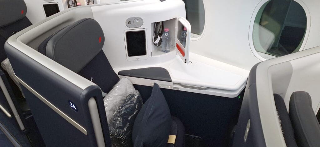 The image shows a business class airplane seat with a sleek, modern design. The seat is dark-colored with a headrest and is positioned next to a window. There is a small storage compartment with a bottle of water and headphones. A pillow and blanket are placed on the seat. The seat number "3A" is visible on the side panel.