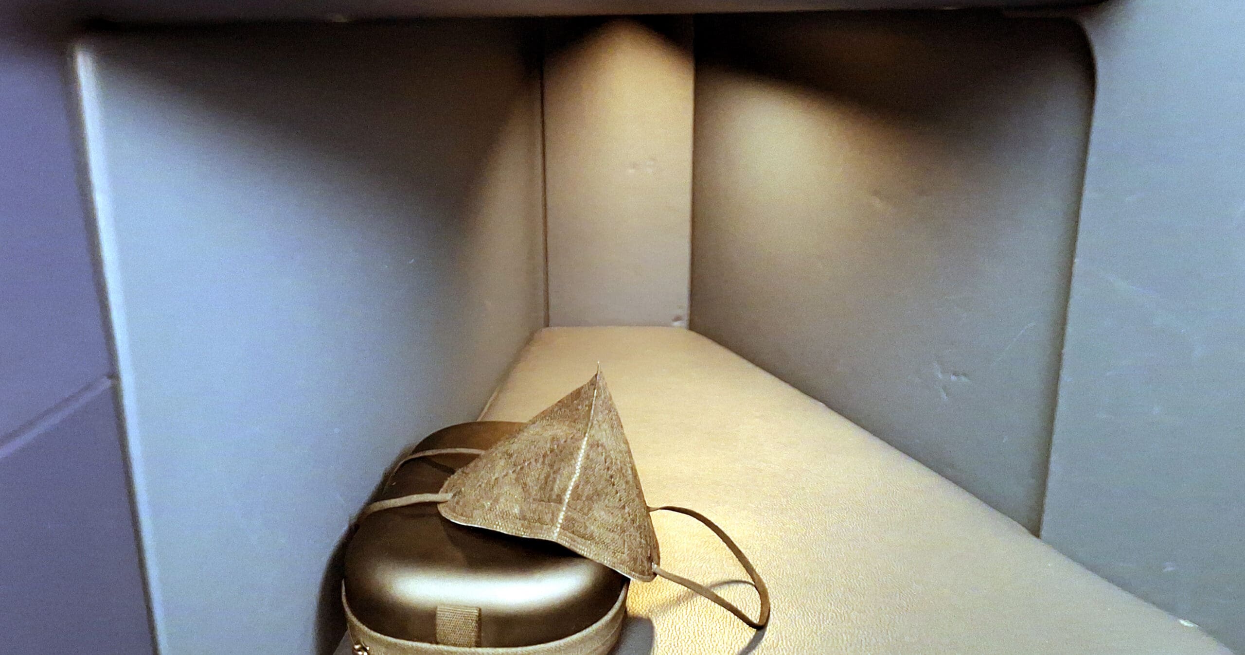 The image shows a small, enclosed storage compartment with a beige interior. Inside, there is a brown, triangular-shaped face mask resting on top of a closed, hard-shell case. The compartment is lit from above, casting soft shadows.