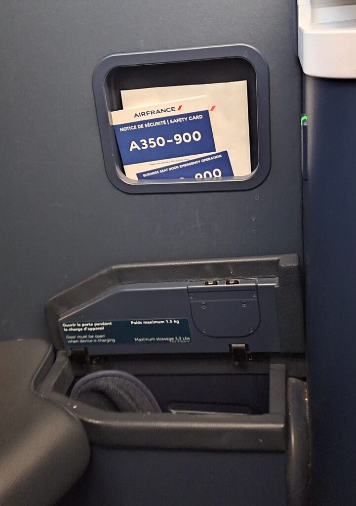 The image shows a section of an airplane interior, specifically a seat area. There is a safety card for an Air France A350-900 aircraft visible in a pocket. Below the pocket, there is a storage compartment with a label indicating a maximum weight limit of 1.5 kg (3.3 lbs). A rolled-up item, possibly a blanket or pillow, is stored in the compartment.