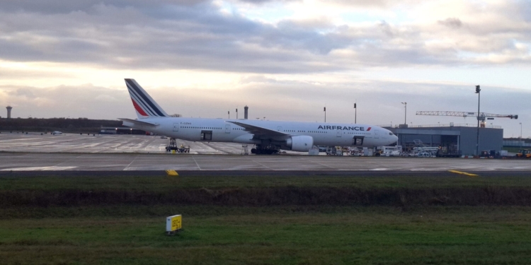 An Air France airplane is parked on the tarmac at an airport. The aircraft is positioned near a terminal building with various ground service vehicles and equipment nearby. The sky is cloudy, and there is a control tower visible in the background.