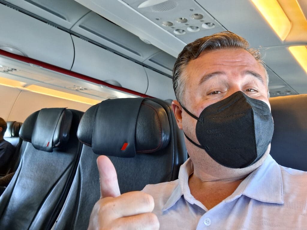 A person wearing a black face mask is sitting in an airplane seat, giving a thumbs-up gesture. The airplane cabin has empty seats with black leather headrests visible in the background. The person is wearing a light-colored shirt.