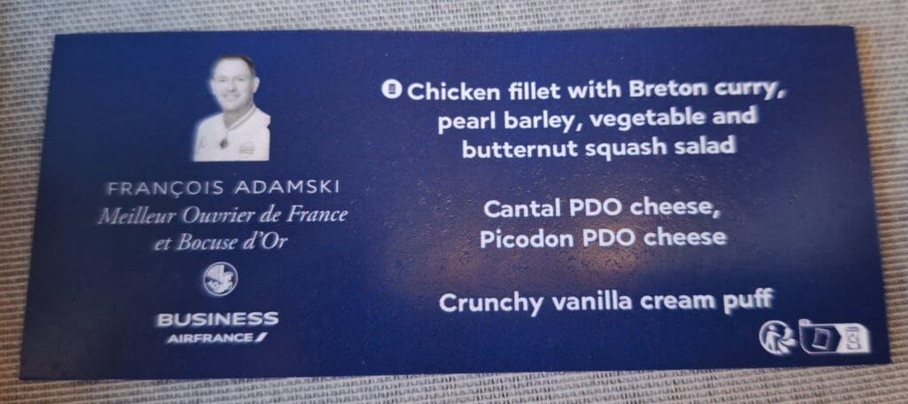 The image shows a blue menu card with a small black-and-white photo of a person. The text on the card includes: - "Chicken fillet with Breton curry, pearl barley, vegetable and butternut squash salad" - "Cantal PDO cheese, Picodon PDO cheese" - "Crunchy vanilla cream puff" Below the photo, it reads: - "FRANÇOIS ADAMSKI" - "Meilleur Ouvrier de France et Bocuse d'Or" - "BUSINESS AIRFRANCE" with the Air France logo.