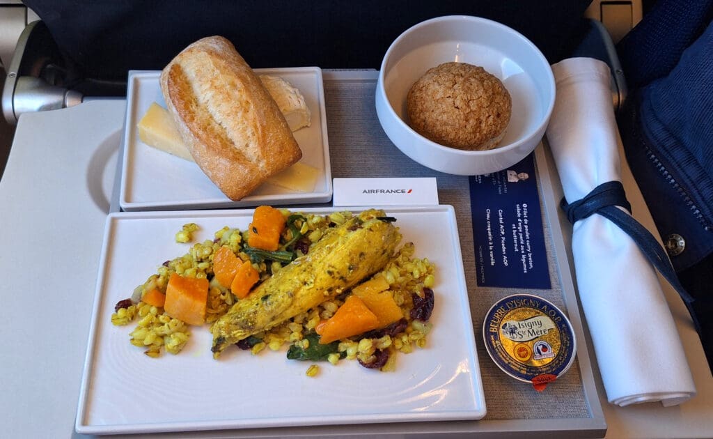 An airplane meal served on a tray, featuring a main dish of seasoned chicken with grains, squash, and vegetables. There is a side plate with a piece of bread and cheese, a small bowl with a cookie, a packet of butter, and a rolled napkin with cutlery. The tray also includes an Air France card.