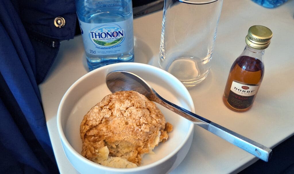 The image shows a bowl with a dessert that appears to be a crumble or cobbler, with a spoon resting inside. Next to the bowl is a small bottle of Thonon water, an empty glass, and a miniature bottle of Dobbé cognac. The items are placed on a light-colored surface, possibly a table or tray.