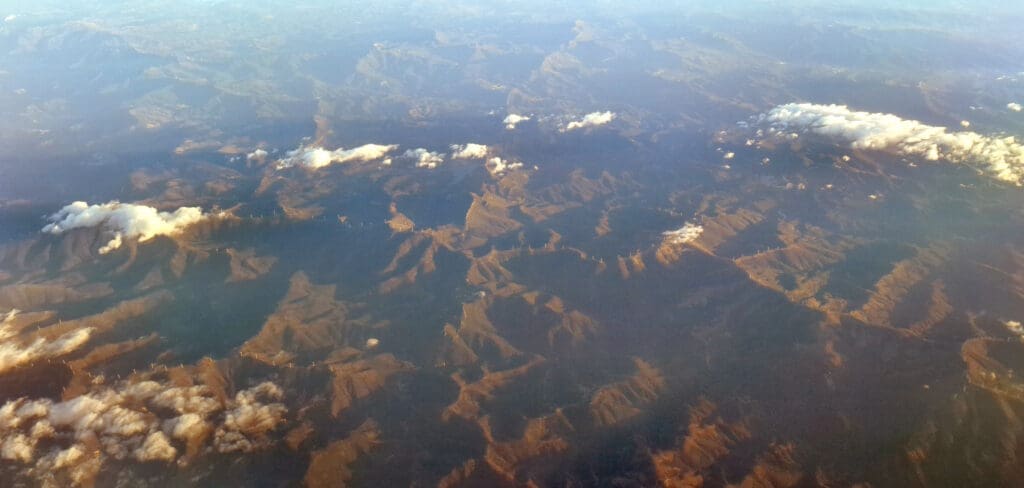 Aerial view of a mountainous landscape with ridges and valleys. The terrain is partially covered by scattered clouds, casting shadows on the ground. The lighting suggests it is either early morning or late afternoon, with a warm glow over the scene.