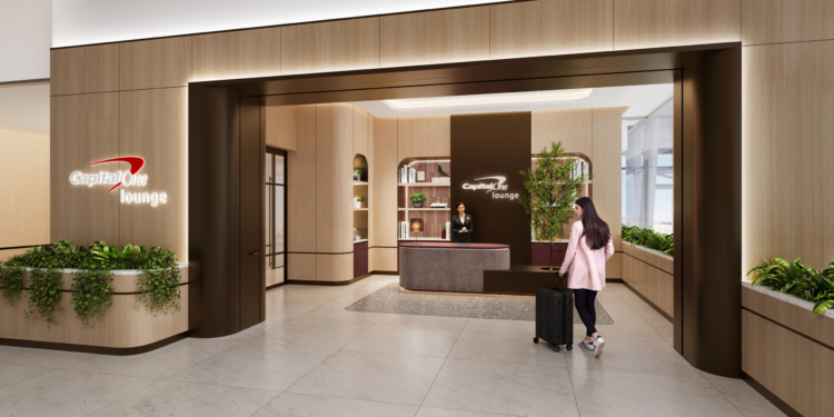 The image shows the entrance to a Capital One Lounge. The design features a modern and elegant interior with wooden paneling and greenery. A person with long hair, wearing a pink coat, is walking towards the reception desk, pulling a suitcase. A staff member is standing behind the desk, and the lounge's logo is visible on the wall.