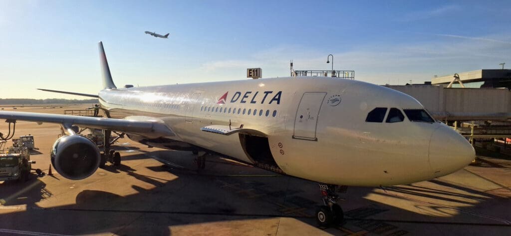 A large commercial airplane is parked at an airport gate, with the Delta logo visible on its side. The aircraft is connected to a jet bridge, and there are ground service vehicles nearby. In the background, another airplane is taking off against a clear blue sky.