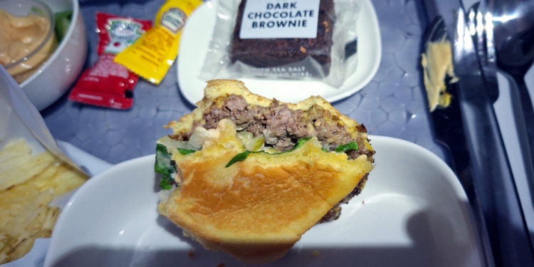 A partially eaten hamburger is on a white plate, showing layers of beef, lettuce, and sauce. Nearby, there are packets of ketchup and mustard, a bowl with a sauce, a bag of chips, and a packaged dark chocolate brownie. A fork and knife are on the tray.