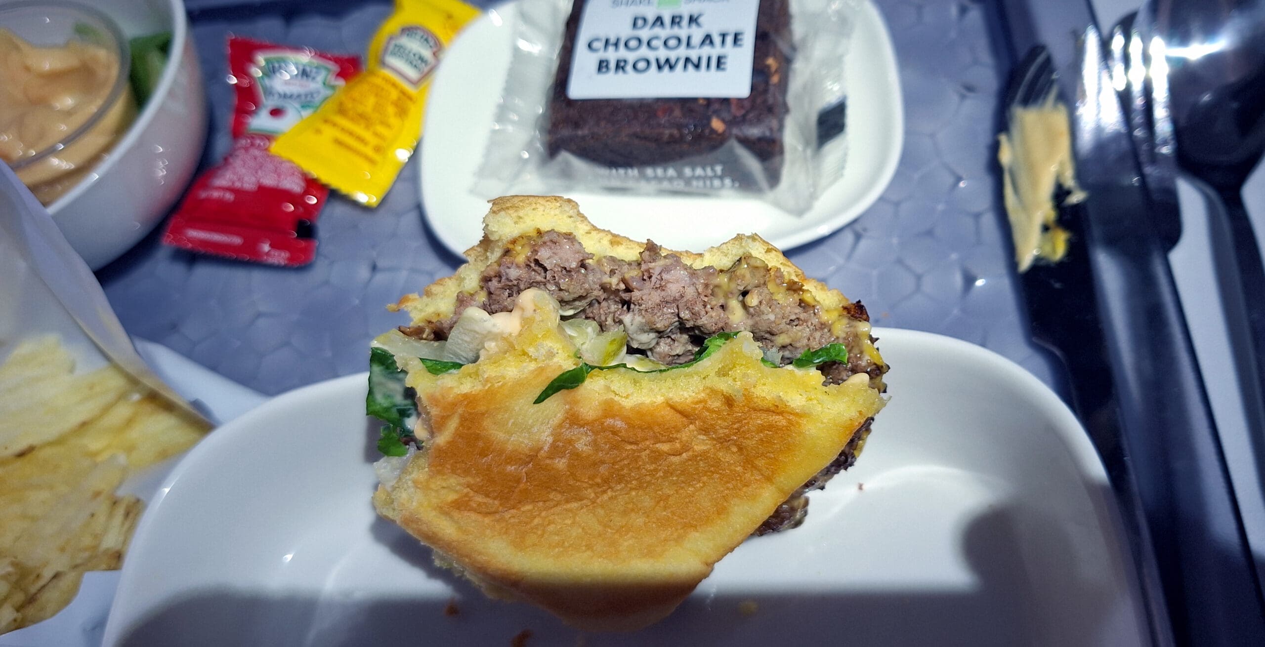 A partially eaten hamburger is on a white plate, showing layers of beef, lettuce, and sauce. Nearby, there are packets of ketchup and mustard, a bowl with a sauce, a bag of chips, and a packaged dark chocolate brownie. A fork and knife are on the tray.
