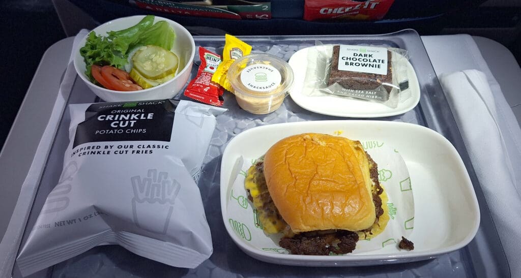 An airplane meal tray featuring a cheeseburger in a bun, a small bowl of salad with lettuce, tomato, and pickles, a packet of crinkle-cut potato chips, a dark chocolate brownie, and condiments including ketchup, mustard, and ShackSauce. A napkin is placed on the side.