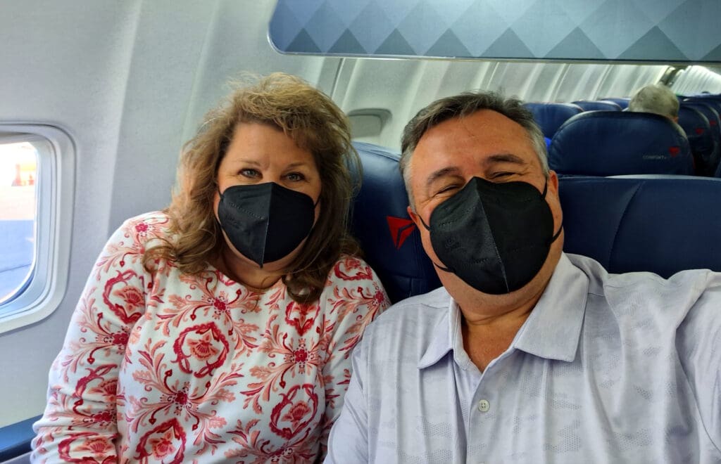 A man and a woman are sitting next to each other on an airplane, wearing black face masks. The woman is wearing a white and red floral patterned top, and the man is wearing a light-colored shirt. They are smiling and appear to be taking a selfie. The airplane seats and window are visible in the background.