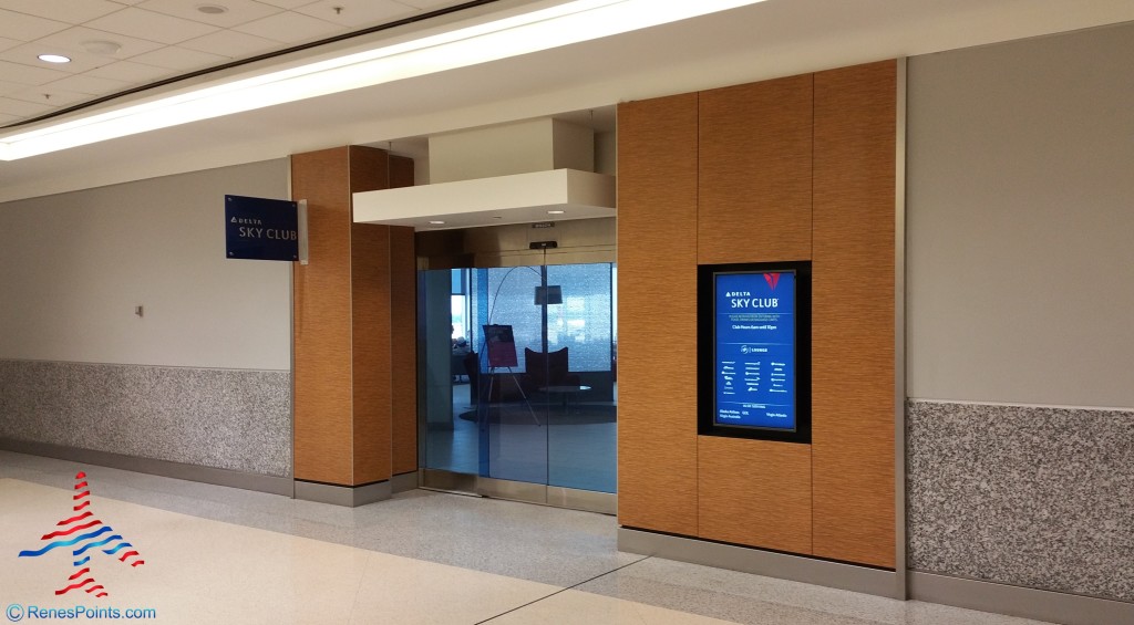 The image shows the entrance to a Delta Sky Club lounge at an airport. The entrance features glass doors and is flanked by wooden panels. There is a digital sign on the right side displaying information about the lounge. The area is well-lit with a clean, modern design.