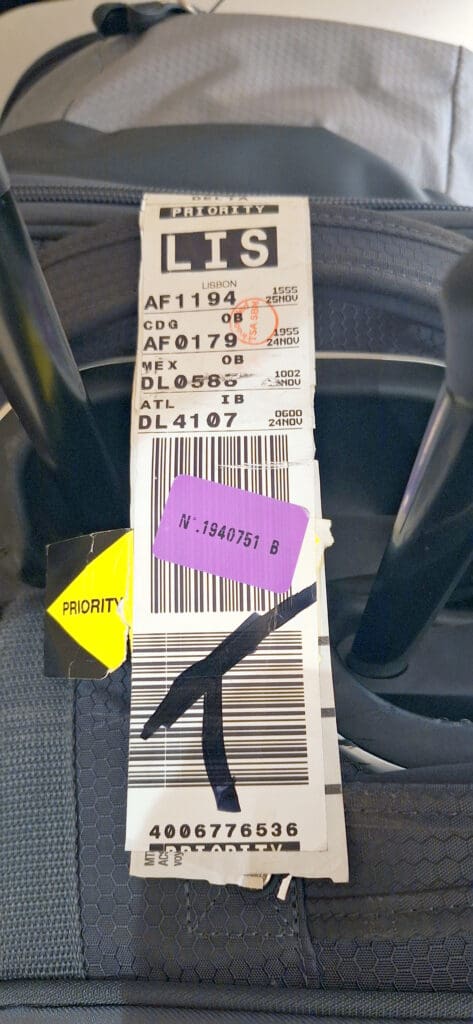 The image shows a close-up of a luggage tag attached to a suitcase handle. The tag has "LIS" in large letters, indicating Lisbon as the destination. It includes flight details such as AF1194, AF0179, DL0588, and DL4107, with corresponding airport codes CDG, MEX, and ATL. The tag is marked "PRIORITY" and has a barcode and a purple sticker with the number "N: 1940751 B." The suitcase is dark-colored with a textured fabric.