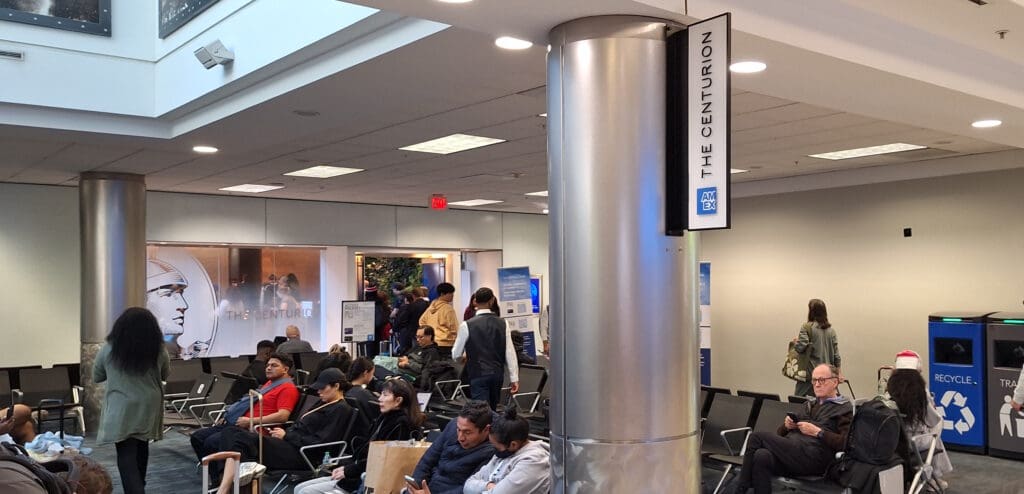 The image shows a busy airport lounge area with several people seated and waiting. There is a sign on a pillar that reads "The Centurion" with the AMEX logo. In the background, there is a glass entrance with the same name and a large image of a centurion. People are seen walking and standing around, and there are recycling and trash bins on the right side. The ceiling has recessed lighting, and the area appears to be well-lit.