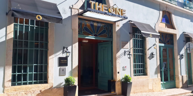 The image shows the exterior of a building with a sign that reads "THE ONE" above the entrance. The facade is light-colored with large windows and decorative iron bars. There are black awnings above the windows and entrance, and two potted plants flank the doorway. The building has a classic architectural style with stone accents and a stained glass design above the door.
