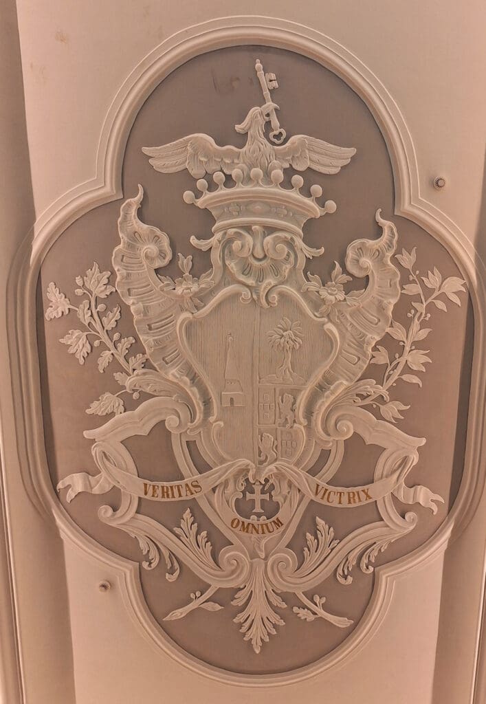 The image shows an ornate, embossed coat of arms on a wall. It features a central shield with intricate designs, including a tree and a building. Above the shield is a crown topped with an eagle. Surrounding the shield are decorative elements like leaves and scrolls. A banner at the bottom displays the Latin words "VERITAS OMNIUM VICTRIX." The entire design is set within an oval frame.