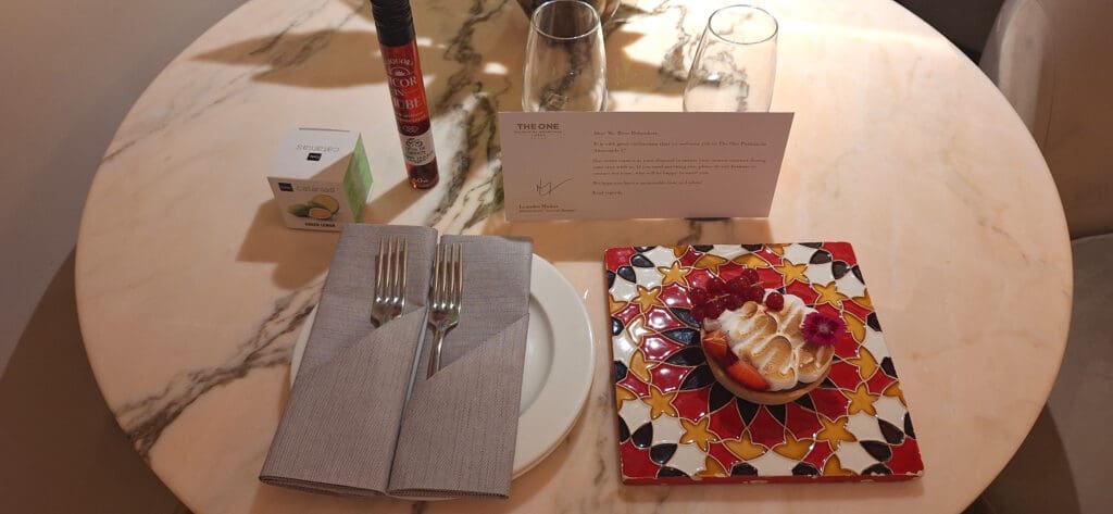 A marble table set with a dessert on a colorful tile, two forks wrapped in napkins on a plate, a small box labeled "Catanias Green Lemon," a tube of liqueur, two empty glasses, and a welcome card from a hotel.