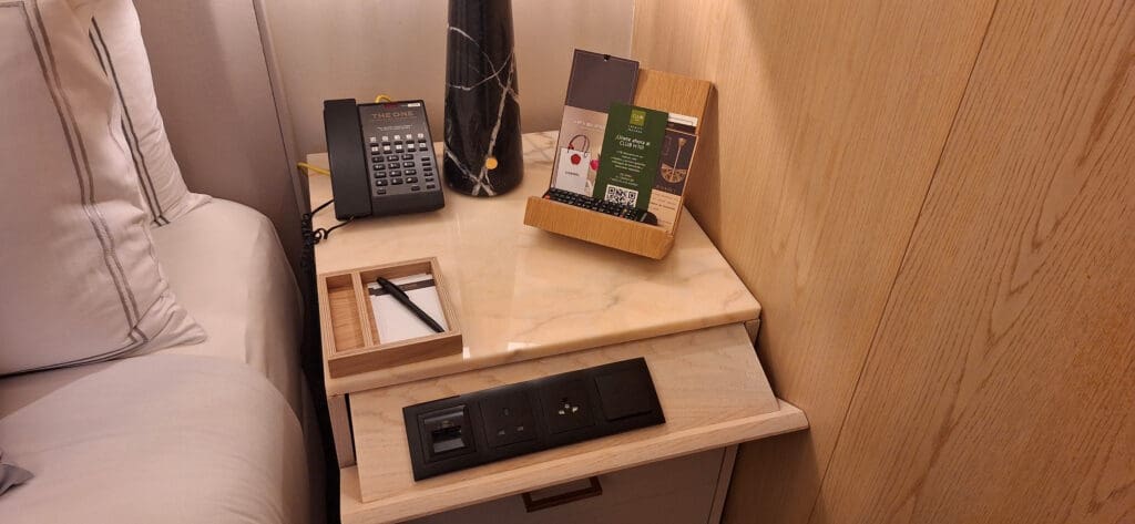 A hotel room nightstand with a marble top. On it, there is a black telephone, a black and white marble-patterned lamp, and a wooden holder containing brochures and a TV remote. A notepad and pen are placed in a wooden tray. Below the tabletop, there are power outlets and USB ports. The bed with white pillows is partially visible on the left.
