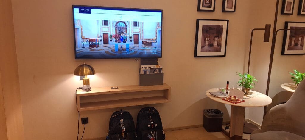 The image shows a cozy hotel room corner with a wall-mounted TV displaying a welcome screen. Below the TV is a small wooden shelf with a modern lamp and a magazine holder containing magazines. Two backpacks are placed on the floor beneath the shelf. To the right, there is a round table with a potted plant, a dessert on a plate, a glass, and some papers. The wall is decorated with framed photographs.