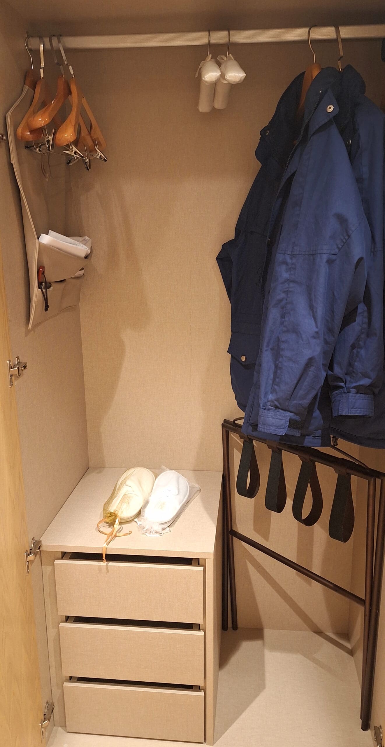 The image shows an open closet with a beige interior. Inside, there are several wooden hangers on a rod, a blue jacket hanging, and two white items hanging from the rod. On the left, there is a fabric organizer with a pair of scissors and some papers. Below, there is a small set of three drawers with two pairs of slippers on top, one white and one beige. A luggage rack with black straps is also visible.