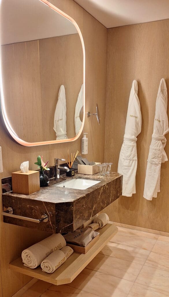 A modern bathroom with a wooden interior features a large, illuminated mirror above a dark marble sink. On the sink are a tissue box, a small plant, a soap dispenser, and a set of toiletries. Two white bathrobes hang on the wall, and below the sink, there are rolled towels and a tray with additional items.