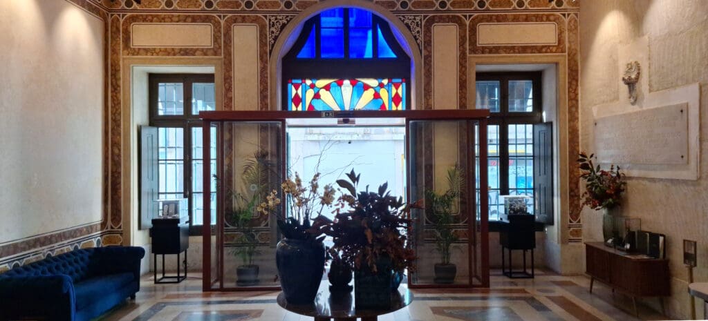 The image shows an elegant interior space with a decorative stained glass window above a set of glass doors. The walls are adorned with intricate patterns and a plaque. There is a blue velvet sofa on the left and a wooden cabinet with decorative items on the right. In the center, there is a round table with large vases filled with plants and flowers. The floor has a geometric pattern.