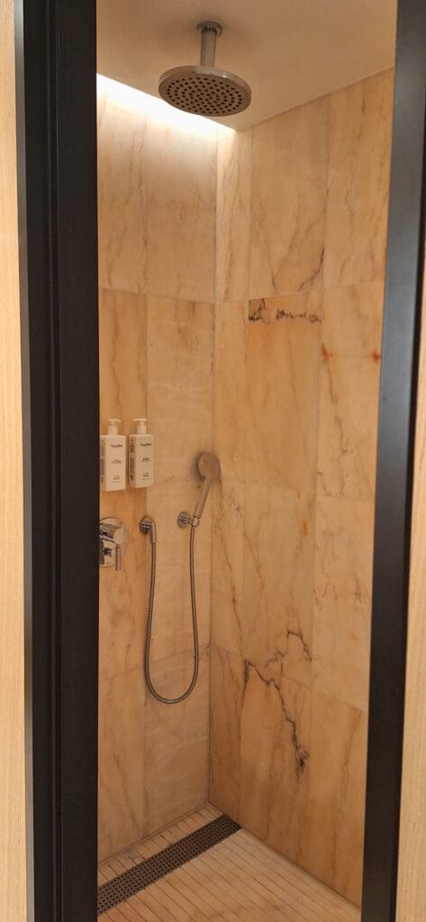A modern shower with beige marble tiles, featuring a ceiling-mounted rain showerhead and a handheld showerhead attached to the wall. There are two wall-mounted bottles for toiletries and a metal drain on the floor.