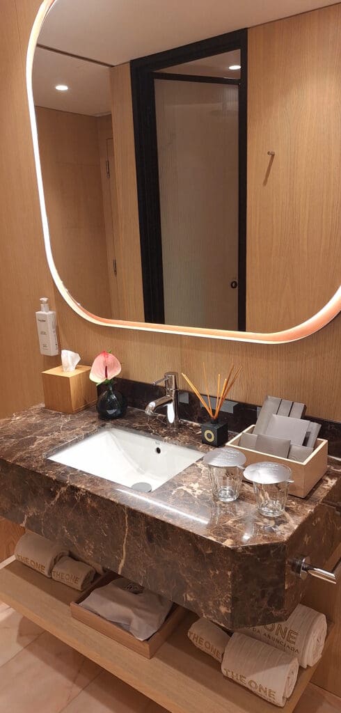 The image shows a modern bathroom vanity area. It features a large, illuminated mirror with a wooden frame. Below the mirror is a marble countertop with a built-in sink. On the countertop, there is a tissue box, a small vase with a pink flower, a set of reed diffusers, and a wooden box containing toiletries. Two glass cups with lids are also present. Below the countertop, there are neatly rolled towels on a wooden shelf. The walls are made of light wood, and a door with a frosted glass panel is visible in the background.