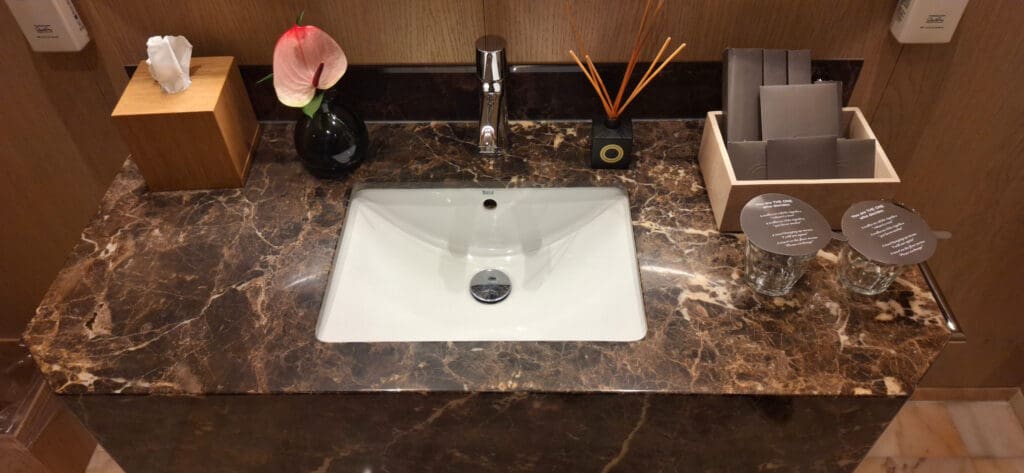 A bathroom countertop with a marble finish features a white sink and a chrome faucet. To the left, there is a wooden tissue box and a black vase with a pink anthurium flower. In the center, a small black container holds orange reed diffusers. On the right, a wooden holder contains several gray paper items. Two glasses with lids are placed on the far right, each with text printed on the lid.