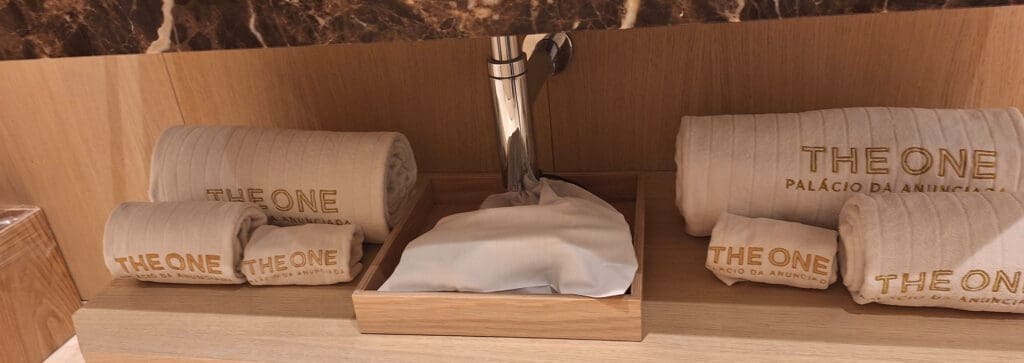 The image shows a set of neatly rolled beige towels with "THE ONE PALÁCIO DA ANUNCIADA" embroidered on them in gold lettering. The towels are placed on a wooden shelf, and there is a wooden tray with a white cloth or cover in the center. The background features a marble surface.
