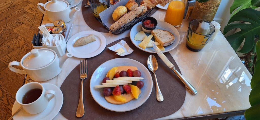 A breakfast table setting with a variety of items. There is a plate of sliced fruits including strawberries, grapes, and melon. Another plate has slices of cheese and bread. A basket contains different types of bread and pastries. There are two teapots, a cup of tea, and a glass of orange juice. Small jars of jam and a dish with butter are also present. The table is set with cutlery and napkins, and there is a plant in the background.
