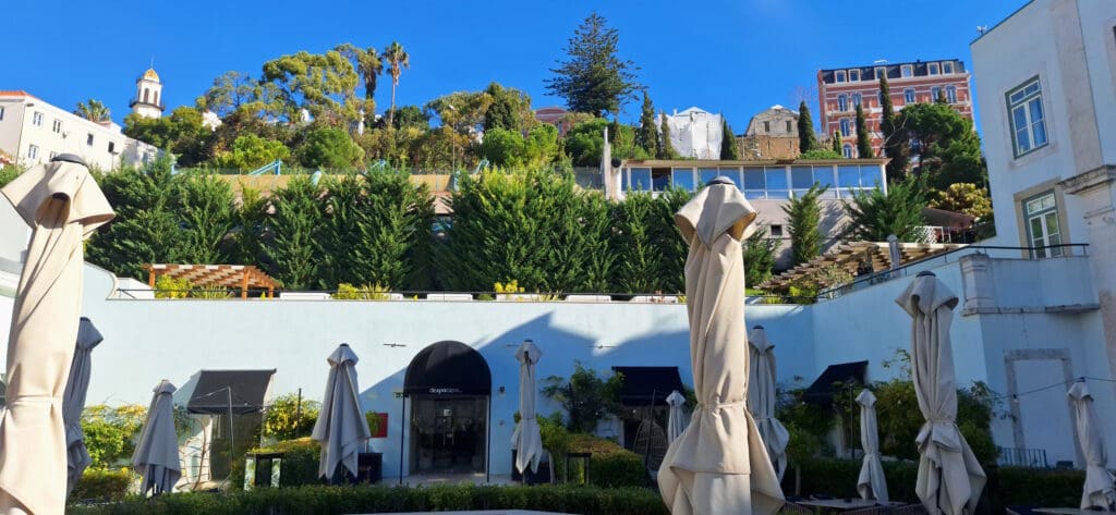 The image shows an outdoor courtyard area with several closed beige umbrellas and tables. In the background, there is a lush hillside with various trees and plants. A building with a dome and another multi-story building are visible at the top of the hill. The sky is clear and blue.