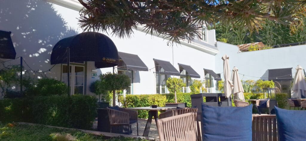 The image shows an outdoor patio area of a restaurant or café. There are several tables and chairs with blue cushions, surrounded by greenery and potted plants. The building has a white exterior with black awnings over the windows. There are also large umbrellas providing shade, and string lights are visible, suggesting a cozy atmosphere.