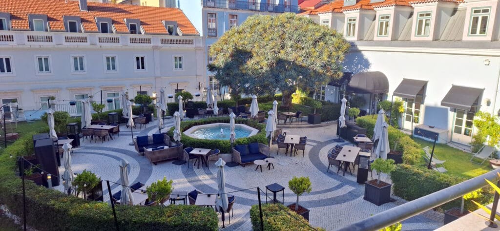 A courtyard with outdoor seating, featuring tables and chairs arranged around a central fountain. The area is surrounded by hedges and trees, with several closed umbrellas near the tables. The courtyard is adjacent to a building with multiple windows and a red-tiled roof.