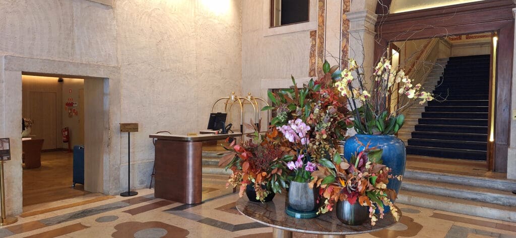 The image shows a hotel lobby with a reception desk on the left and a large floral arrangement on a round table in the center. The flowers are in various colors, including pink, yellow, and orange, placed in blue and gray vases. Behind the desk, there is a luggage cart. To the right, a staircase with dark carpeting leads upward. The walls are decorated with textured patterns and marble accents.