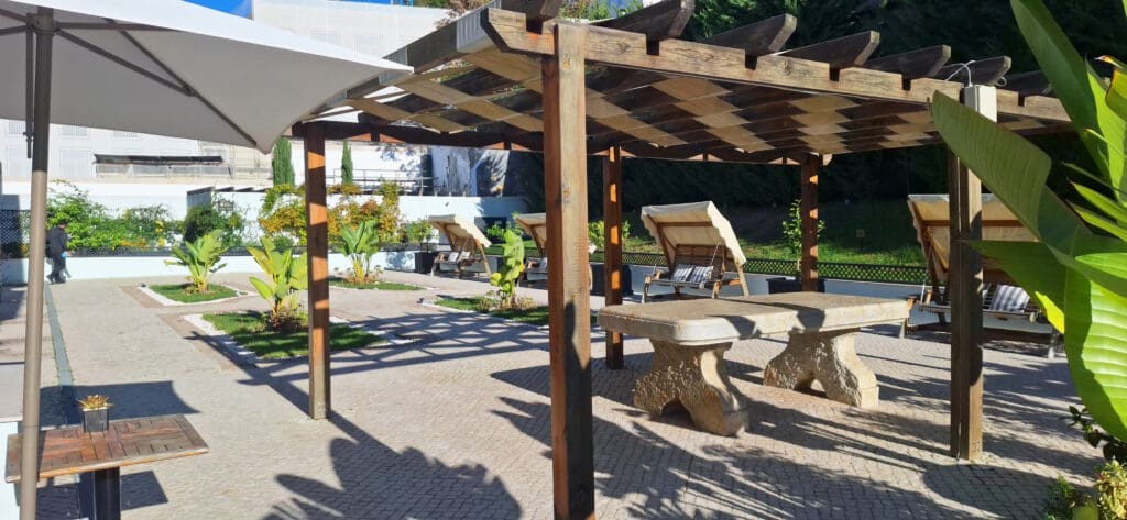 The image shows an outdoor patio area with a wooden pergola providing shade. Underneath the pergola, there are stone tables and benches. The area is surrounded by greenery, including potted plants and small trees. In the background, there are lounge chairs with striped cushions. A large umbrella provides additional shade on the left side of the image. The setting appears to be a garden or a recreational outdoor space.