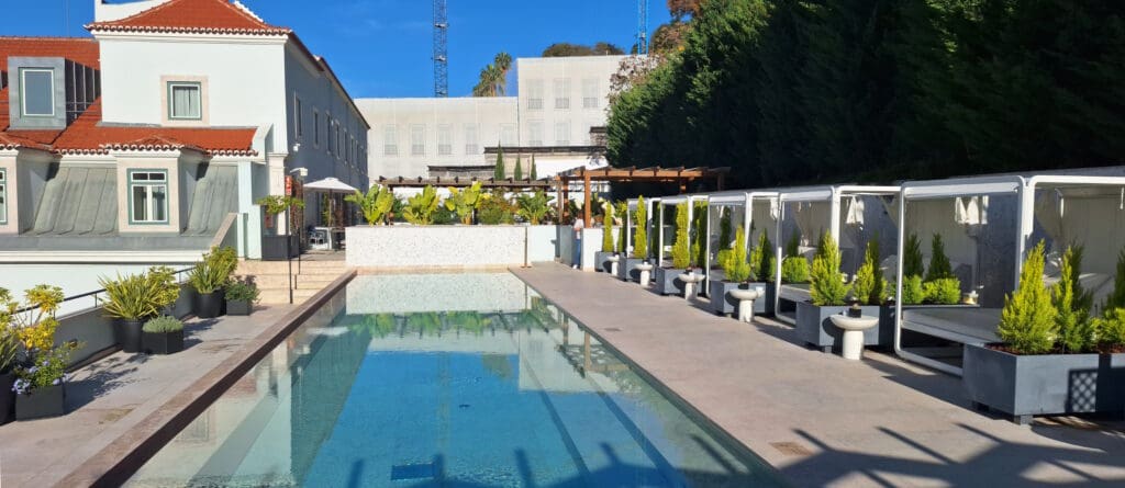 A luxurious outdoor pool area with a clear blue swimming pool surrounded by a modern building with red-tiled roofs. On the right, there are several cabanas with white curtains and potted plants. The left side features more plants in pots and a staircase leading to a patio with tables and umbrellas. The background shows a tall hedge and a clear blue sky.
