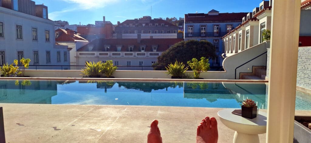 A rooftop pool with clear blue water is surrounded by buildings with traditional architecture. In the foreground, a person’s feet are visible, suggesting they are relaxing by the pool. There are potted plants along the pool's edge, and a small table with a plant on it. The sky is clear and sunny.