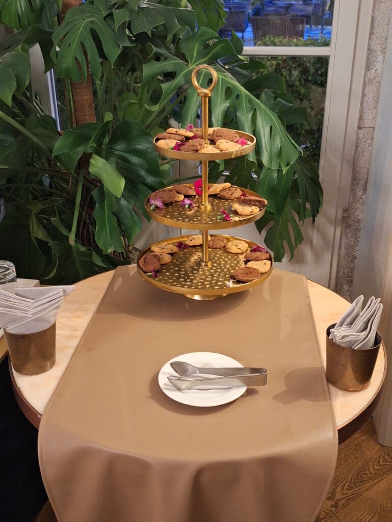 The image shows a three-tiered gold serving stand on a round table with a beige tablecloth. Each tier is filled with cookies and decorated with small pink flowers. The table is set with a white plate and metal tongs. There are napkins in a holder on the table. In the background, there are large green leaves from a plant, and a window showing an outdoor view.