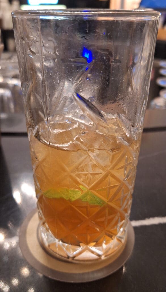 A close-up of a textured glass filled with an amber-colored drink, ice cubes, and a slice of lime. The glass is placed on a coaster on a dark surface, with a blurred background.